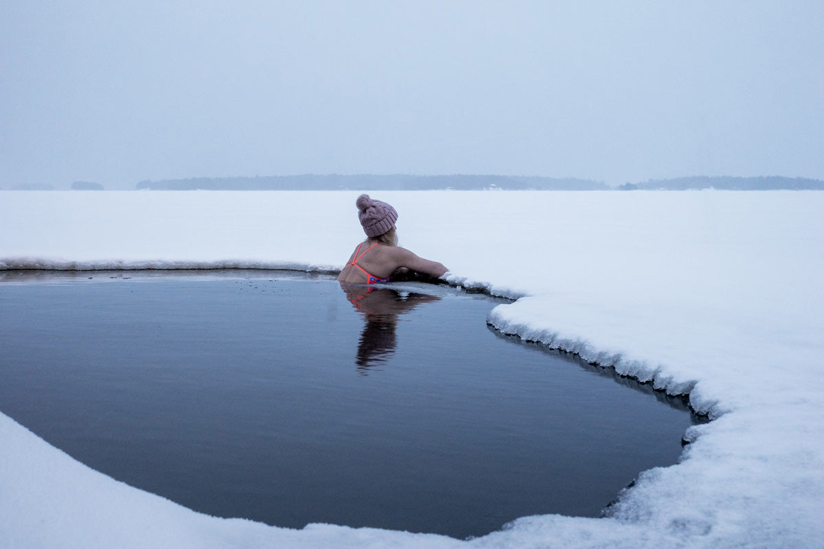 Frau mit Bikini und Pudelmütze badet im Wasser eines Eislochs